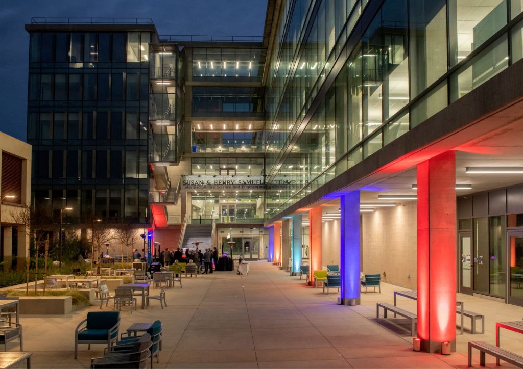 The exterior of the ISEB after dark, with some columns lit in red and blue colored lights. The building's name is visible in the back of the photo in letters mounted to the building.